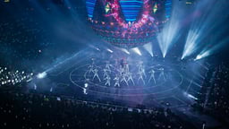 Esiintyjiä lavalla suuressa musiikkikilpailussa, näyttävä valoshow ja yleisö areenalla.
