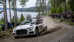 Ralliauto ajaa soratiellä metsämaisemissa yleisön seuratessa erikoiskoetta tien varrella.